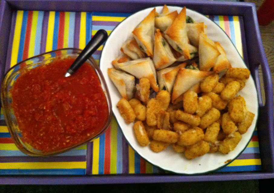 A multi-coloured tray holds two dishes. One holds a homemade marinara sauce (a spoon set into it), and the other holds two types of hors d'oeuvres: spanikopita and mini mozzarella sticks.